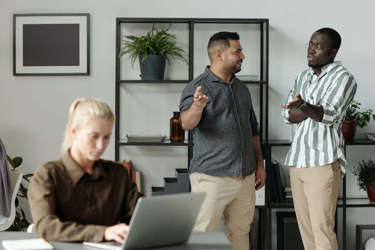 Trois collègues dans un bureau moderne : deux hommes discutent debout devant une étagère, tandis qu’une femme travaille sur un ordinateur portable au premier plan.