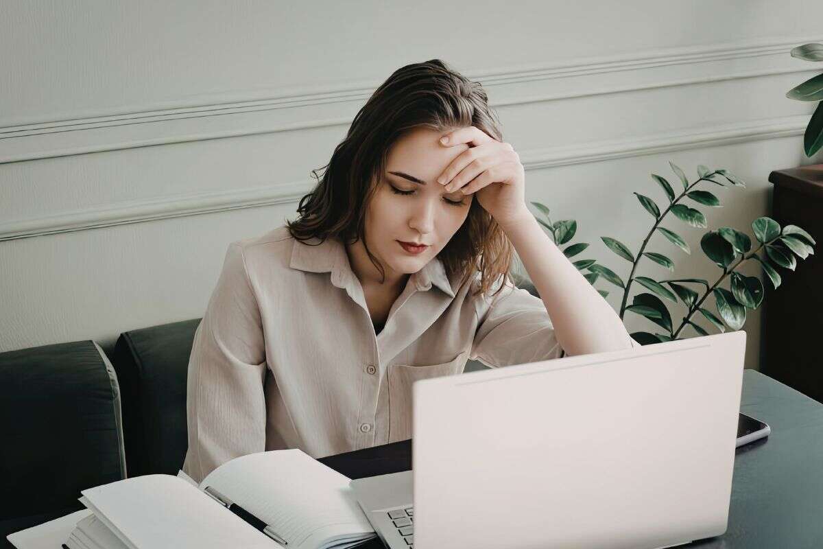 Femme assise à une table, travaillant sur un ordinateur portable, la main sur le front avec une expression de fatigue ou de concentration, entourée d’un carnet et de plantes d’intérieur.