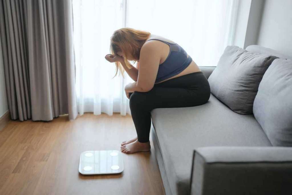 une femme ronde est assise sur son canapé et se tient la tête, elle paraît triste