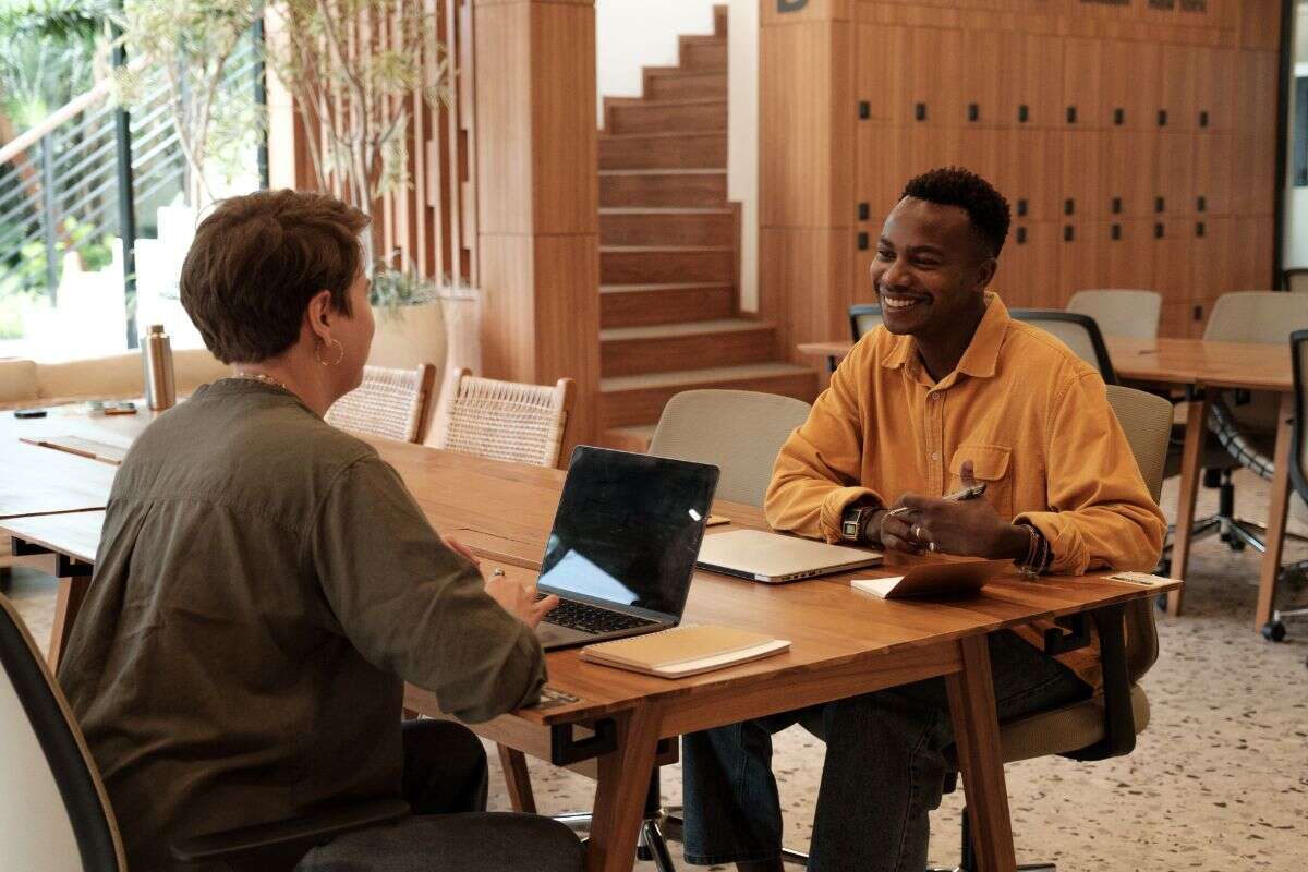Deux personnes échangent autour d’un ordinateur portable dans un espace de travail partagé lumineux, avec tables en bois, casiers et escalier en arrière-plan.